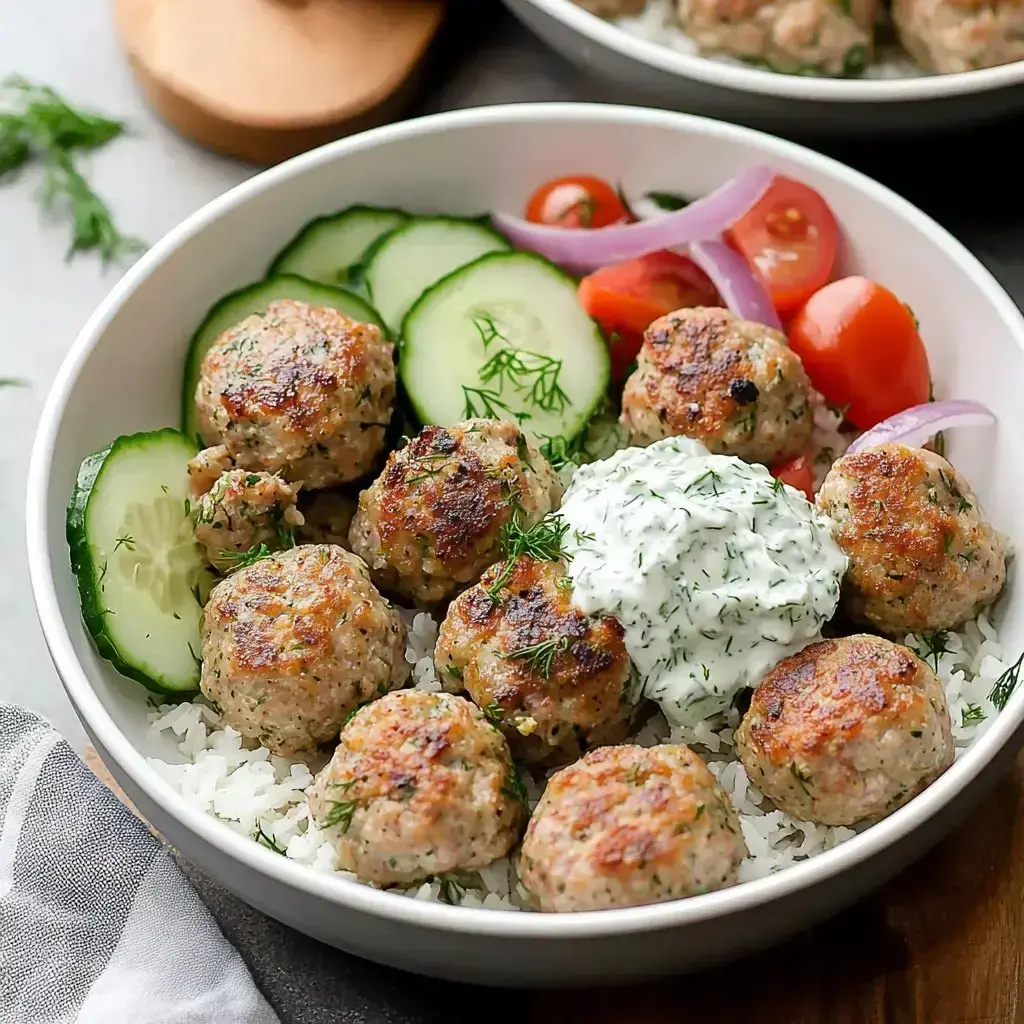 A bowl filled with rice topped with golden-brown meatballs, sliced cucumbers, cherry tomatoes, red onions, and a dollop of creamy herb sauce.