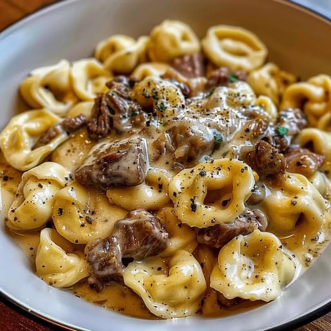 Un bol de pâtes tortellinis nappées de sauce crémeuse avec morceaux de viande et herbes aromatiques.