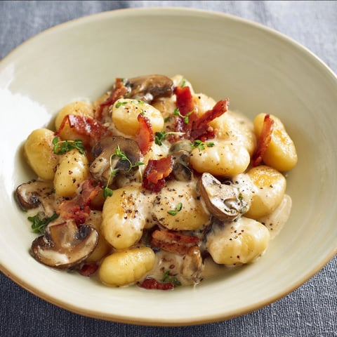 Une grande assiette de gnocchis nappés de fromage coulant avec des champignons poêlés et du bacon grillé.
