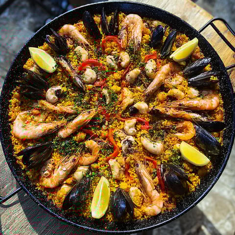 Grande paella de fruits de mer dehors, pleine de langoustines, de poivrons, d’oignons et de tomates, sur une planche en bois.